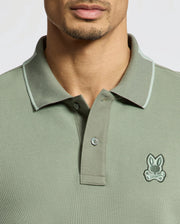 Someone is wearing a Psycho Bunny MENS NEW CASTLE PIQUE POLO - B6K487F200 in green, made of premium Pima Cotton. It features a small embroidered stylized animal logo on the chest, with a collar and button placket on a plain, light background.