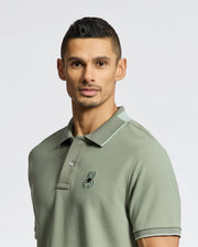 A person stands against a plain white background wearing a soft Pima Cotton Men's New Castle Pique Polo (B6K487F200) by Psycho Bunny in green, featuring a small embroidered logo on the chest.