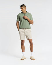 A person stands confidently in a light green Psycho Bunny MENS NEW CASTLE PIQUE POLO - B6K487F200 and beige shorts, hands in pockets, paired with white socks and luxury-finished beige sneakers against a plain white background.