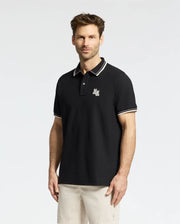 A man with short brown hair and a beard wears the Psycho Bunny MENS ELLIOT PIQUE POLO (B6K505F200), a black Pima cotton polo with white trim and an embroidered logo on the chest. He pairs it with beige shorts against a plain, light-colored background.