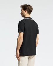 A man with short brown hair, wearing a Psycho Bunny MENS ELLIOT PIQUE POLO - B6K505F200 in black Pima cotton with white trim, faces away from the camera against a plain white background, flawlessly paired with beige pants.