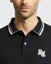 A man sports a Psycho Bunny MENS ELLIOT PIQUE POLO - B6K505F200 in black with a striking white and black striped collar. Crafted from Pima cotton for a perfect fit, it features an embroidered logo of two cartoon rabbits on the chest, set against a plain white background.
