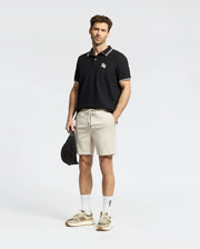 A man exudes confidence in his black MENS ELLIOT PIQUE POLO by Psycho Bunny, featuring bold design elements and a subtle logo. Paired with beige shorts, white socks, and sneakers, his stylish appearance is enhanced by neatly styled hair and a light beard against the plain white backdrop.