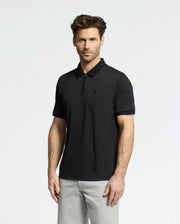 A man with short brown hair and a beard wears the Psycho Bunny MENS TREVOR PERFORMANCE PIQUE ZIP UP POLO (B6K557F200) in black and light gray pants, standing against a plain white background.