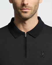 A man wearing the Psycho Bunny MENS TREVOR PERFORMANCE PIQUE ZIP UP POLO in black, featuring a slim fit, collared design, front zipper, and an embroidered bunny and crossbones logo on the left chest. His face is shown from shoulders to above the lips.