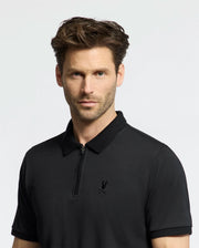A man with short brown hair and light stubble wears the Psycho Bunny MENS TREVOR PERFORMANCE PIQUE ZIP UP POLO (B6K557F200), a black slim-fit collared shirt with an embroidered bird logo, standing against a plain light background.
