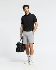 A man stands against a plain background wearing the Psycho Bunny MENS TREVOR PERFORMANCE PIQUE ZIP UP POLO in black, light gray shorts, white socks with a black logo, white sneakers, and holding a black duffel bag by his side.