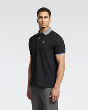 A man with short curly hair models the Psycho Bunny Men's Jonas Stripe Collar Pique Polo (B6K583F200), crafted from luxurious Peruvian Pima Cotton. It features a black design, gray collar and cuffs with micro-stripe detail, and an embroidered chest logo. He stands against a plain white background, looking at the camera.
