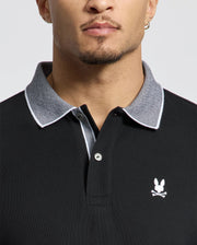 A person wearing a black Psycho Bunny MENS JONAS STRIPE COLLAR PIQUE POLO with a gray collar and small white bunny logo above crossed bones. Made from Pima Cotton, it features micro-stripes. The focus is on the upper torso and neck, showcasing short hair and a stud earring.