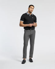 A man stands against a plain background wearing a Psycho Bunny Mens Jonas Stripe Collar Pique Polo in black, featuring a gray collar and cuffs. He pairs it with gray pants and black loafers, holding an item near his waist.
