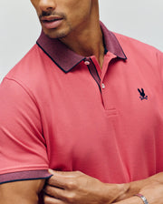 A man crosses his arms wearing the Psycho Bunny MENS JONAS MICRO STRIPE COLLAR PIQUE POLO (B6K583F200) in red, featuring micro-stripe accents, a dark collar, and a small navy bunny logo. Only his lower face and upper torso are visible.