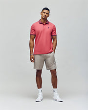 A man with short hair and a neutral expression stands against a plain background wearing the Psycho Bunny MENS JONAS MICRO STRIPE COLLAR PIQUE POLO in pink, paired with beige shorts, white socks, white sneakers, and one hand in his pocket.