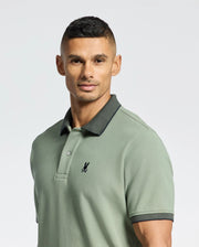A person wears the Psycho Bunny MENS JONAS MICRO STRIPE COLLAR PIQUE POLO, featuring a dark collar and an embroidered chest logo. They stand against a light background with short dark hair and a neutral expression.