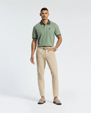 A man in a Psycho Bunny MENS JONAS MICRO STRIPE COLLAR PIQUE POLO, crafted from premium Pima cotton and light green, stands against a plain white background. He has short hair, a watch on his left wrist, and pairs it with beige pants and light brown loafers.