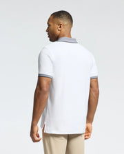 A man stands facing away in a Psycho Bunny MENS JONAS STRIPE COLLAR PIQUE POLO (B6K583F200), featuring white Pima cotton with gray-striped collar and sleeves, paired with beige pants against a plain light background.