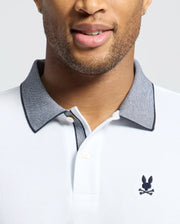 A person wears the Psycho Bunny MENS JONAS STRIPE COLLAR PIQUE POLO (B6K583F200), white with a grey stripe collar and small black bunny skull and crossbones logo on the chest. Only the lower half of the face and upper torso are visible.