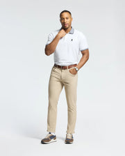 A man poses against a plain background wearing the Psycho Bunny MENS JONAS STRIPE COLLAR PIQUE POLO (B6K583F200), beige pants, a brown belt, watch, and sneakers. He rests one hand on his neck and looks at the camera.