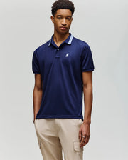 A young man wears the Psycho Bunny MENS FORREST SPORT PIQUE POLO (B6K660F200), a navy blue slim-fit shirt with a white logo, paired with beige cargo pants, standing against a plain, light background.