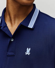 Close-up of a person wearing the Psycho Bunny MENS FORREST SPORT PIQUE POLO (B6K660F200) in dark blue with light blue and white collar trim, breathable moisture-wicking fabric, and a small bunny head above crossed bones on the chest.