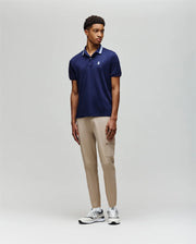 A man stands against a plain background wearing the Psycho Bunny MENS FORREST SPORT PIQUE POLO - B6K660F200, featuring a navy slim-fit design with a light blue collar, paired with beige cargo pants and white sneakers for all-day comfort and style.