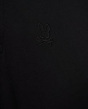 Close-up of the Psycho Bunny MENS JAMES PREMIUM PIQUE POLO SHIRT - B6K945D200 in black, showcasing an embroidered logo depicting a rabbit's head with crossbones underneath. The all-black logo blends subtly with the fabric, adding a refined touch. Made from premium Pima cotton, this durable shirt offers both style and comfort.