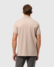 A man with light brown hair is standing with his back to the camera, dressed in a beige MENS JAMES PREMIUM PIQUE POLO SHIRT by Psycho Bunny and dark gray pants. The background is plain and light-colored.