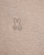 Close-up image of a beige fabric featuring an embroidered logo of a bunny head above two crossed bones, offering a playful twist on the classic skull and crossbones symbol. The embroidery, crafted in a slightly darker shade of beige, enhances the unique character of this durable Psycho Bunny James Premium Pique Polo Shirt - B6K945D200 made from premium Pima cotton.