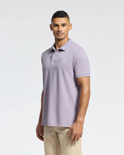 A man in a Psycho Bunny MENS JAMES PIQUE POLO - B6K945D200, a durable Pima cotton light purple shirt, paired with beige pants stands against a white background, looking at the camera with a neutral expression.