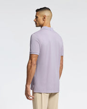 A person stands facing slightly away, dressed in a light purple MENS JAMES PIQUE POLO - B6K945D200 by Psycho Bunny, crafted from durable Pima cotton, paired with beige pants against a plain white background.