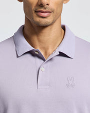 A person is wearing the MENS JAMES PIQUE POLO - B6K945D200 by Psycho Bunny, a Slim Fit light purple polo with a collar, two buttons, and an embroidered bunny with crossbones logo on the left chest. Made from durable Pima cotton. The background is light gray, showing just the lower face.