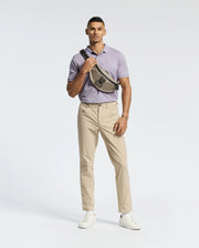 A person confidently wears a slim fit MENS JAMES PIQUE POLO by Psycho Bunny in light purple, paired with beige pants and white sneakers. A black crossbody bag with a bird motif is slung across their chest against a plain white background.