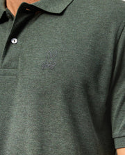 Close-up of a person wearing a dark green Psycho Bunny Men’s James Premium Pique Polo Shirt, model B6K945D200, with two buttons and a small embroidered logo of a rabbit sitting above crossbones on the left chest. The high-quality polo for men features textured fabric, and the image focuses on the upper torso area.