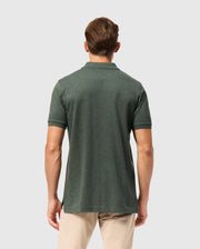 A man is standing against a plain background, facing away from the camera. He is wearing a dark green Psycho Bunny MENS JAMES PREMIUM PIQUE POLO SHIRT - B6K945D200 and beige pants. The polo shirt has short sleeves and a collar.