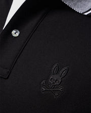 Close-up of the Psycho Bunny Men's Malton Pique Polo Shirt in black Pima cotton, showcasing an embroidered Psycho Bunny skull and crossbones logo on the left chest. The collar features a white and black striped pattern, and the shirt is designed with a button.