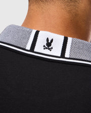 Close-up image of the back of a person's neck, showing the collar of a black and white diamond-knit Pima cotton MENS MALTON PIQUE POLO SHIRT - B6K950D200 by Psycho Bunny. The collar features a small logo of a stylized Psycho Bunny head over crossbones, embroidered in black. The background is plain and light-colored.
