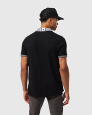 A man wearing a Psycho Bunny MENS MALTON PIQUE POLO SHIRT - B6K950D200 with an emblem on the back of the collar is standing with his back to the camera. He is also wearing dark pants, a black baseball cap, and has short hair. The plain light gray background makes him stand out effortlessly.