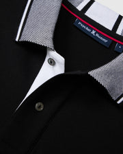 Close-up of a black Pima cotton polo shirt from Psycho Bunny, specifically the MENS MALTON PIQUE POLO SHIRT - B6K950D200. The shirt showcases a textured, diamond-knit fabric with black buttons and a collar adorned with a black and white checkered pattern. A visible tag on the inside neck displays the Psycho Bunny logo and brand name.