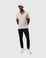 A man stands against a plain background wearing a light grey Psycho Bunny MENS MALTON PIQUE POLO SHIRT - B6K950D200, black pants, white sneakers, and a light grey cap. His hands are clasped behind his back, and he has a relaxed, casual style.