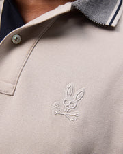 Close-up of a beige Psycho Bunny Malton Pique Polo Shirt (B6K950D200) made from Pima cotton, featuring a small emblem on the chest depicting a rabbit skull and crossbones. The collar is a contrasting grey color with striped detailing and features a subtle diamond pique pattern. Only part of the person's neck and collarbone is visible.