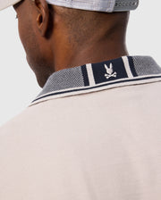 Close-up of the back of a person wearing the light beige MENS MALTON PIQUE POLO SHIRT - B6K950D200 from Psycho Bunny, featuring a navy-collared trim with white stripes and a small Psycho Bunny logo above crossed bones. The shirt is crafted from soft Pima cotton. The person is also wearing a gray cap backward.