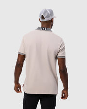 A person is wearing the beige Psycho Bunny MENS MALTON PIQUE POLO SHIRT - B6K950D200, which features black and white detailing on the collar and cuffs, paired with a white baseball cap. The individual, epitomizing the understated elegance of Psycho Bunny, stands facing away from the camera against a plain background.