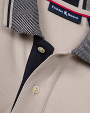 Close-up of a beige Psycho Bunny Malton Pique polo shirt (product code: B6K950D200), featuring a contrasting collar with a black and white checkered pattern. The shirt's placket is partially buttoned with two tan buttons visible, and the collar tag displays the Psycho Bunny logo and brand name.