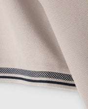 Close-up of the MENS MALTON PIQUE POLO SHIRT - B6K950D200 by Psycho Bunny, featuring a light beige fabric with a textured weave reminiscent of a diamond pique pattern. The edge of the shirt displays a clean, folded hem accented by two thin, parallel black lines running along its length, adding a touch of contrast to the design.