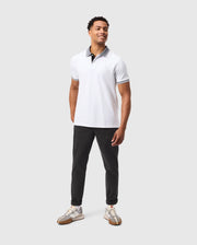 A man stands smiling in the Psycho Bunny Men's Malton Pique Polo Shirt—B6K950D200, featuring a contrasting collar paired with dark trousers. His curly hair adds charm, and he's sporting sneakers. The plain gray background makes him the focus of this Psycho Bunny-inspired look.