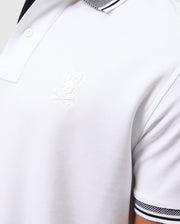 A close-up of a person wearing the MENS MALTON PIQUE POLO SHIRT - B6K950D200 by Psycho Bunny showcases a white Pima cotton polo with black and white striped collar and sleeve accents. The shirt is adorned with an embroidered Psycho Bunny logo depicting a bunny head with crossbones on the chest, all crafted from exquisite fine diamond knit fabric for an added layer of sophistication.