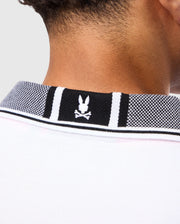Close-up of the back of a person's neck wearing a Psycho Bunny MENS MALTON PIQUE POLO SHIRT - B6K950D200 in white Pima cotton, featuring a black and white checkered collar. The collar showcases the brand’s iconic logo: a skull and crossbones with bunny ears.