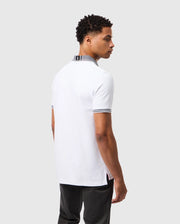 A person with short curly hair is wearing the Psycho Bunny MENS MALTON PIQUE POLO SHIRT - B6K950D200, which features a white Pima cotton design with striped collar and sleeve detailing, facing slightly away from the camera. The plain gray background subtly highlights the distinctive style of Psycho Bunny.
