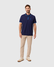 A man stands wearing the stylish Psycho Bunny Men's Kent Pique Polo Shirt, B6K951D200, in navy with blue accents, paired with beige pants and sneakers against a light gray background.