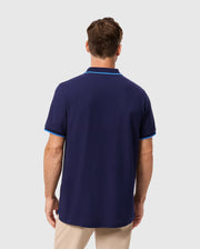 A person wearing a fashionable Psycho Bunny MENS KENT PIQUE POLO SHIRT in dark blue with light blue trim faces away, expertly paired with light khaki pants. The plain gray background highlights the outfit's simplicity and style.