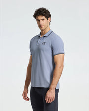 A curly-haired man wears a Psycho Bunny MENS MONROE PIQUE POLO (B6K964D200) in light blue with a subtle chest logo, paired with dark pants. He stands against a plain white background.